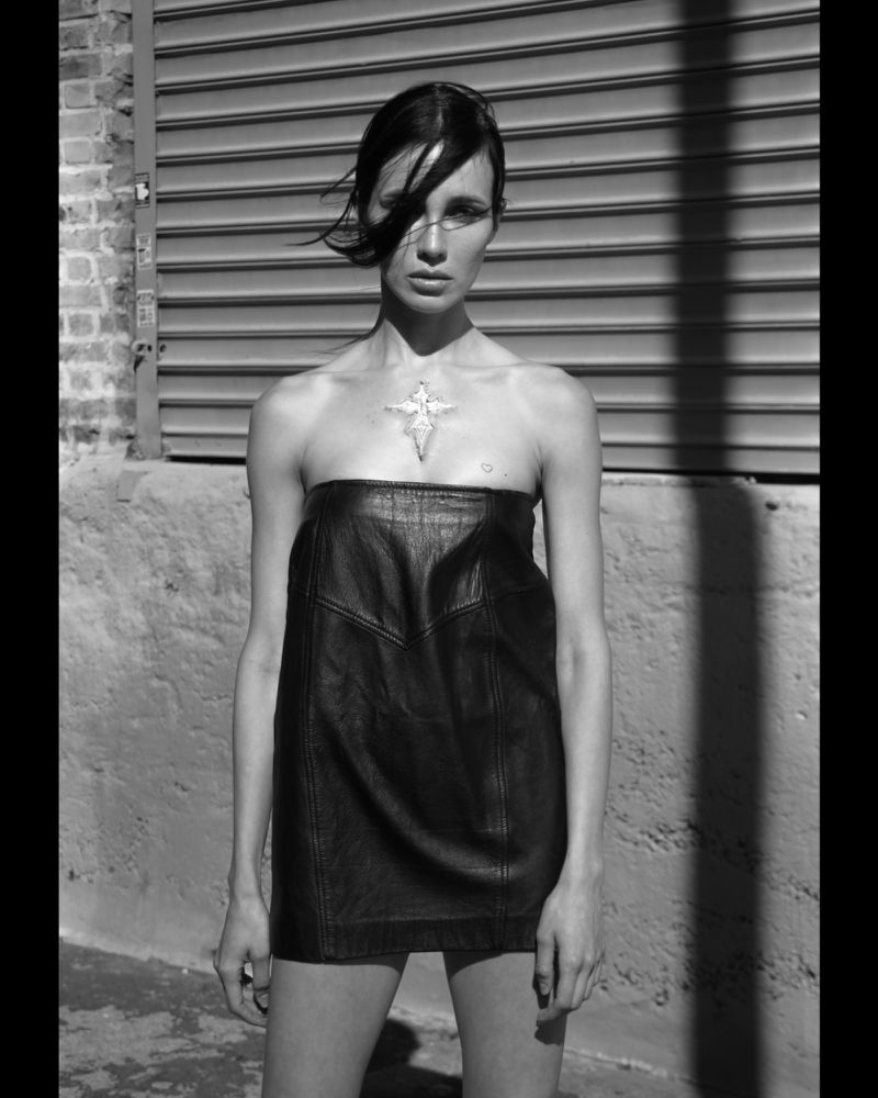 Black-and-white fashion shot of an adult woman wearing a strapless black leather dress, standing outdoors in an urban setting with a concrete wall.