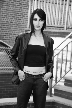 Monochrome portrait of a stylish woman in a blazer and jeans with visible tattoo, posed against a textured brick wall and stairs.