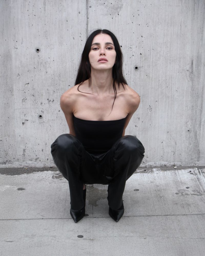 Woman in black tube top and shiny pants squats in front of concrete wall, looking at the camera, fashion photography.