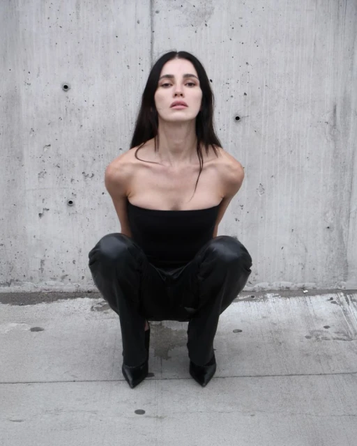 Woman in black tube top and shiny pants squats in front of concrete wall, looking at the camera, fashion photography.