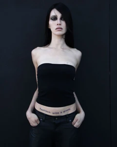 Model with dark eye makeup wearing a black strapless top and black pants against a dark background