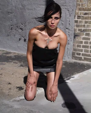 Model kneeling on concrete wearing a black leather dress and pendant against a textured brick wall.