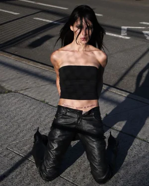 Model with long dark hair wearing a black bandeau top and black leather pants kneeling on an asphalt street.