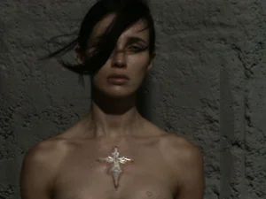 Portrait of a woman with dark hair blowing across her face and a decorative cross emblem on her chest against a textured concrete wall