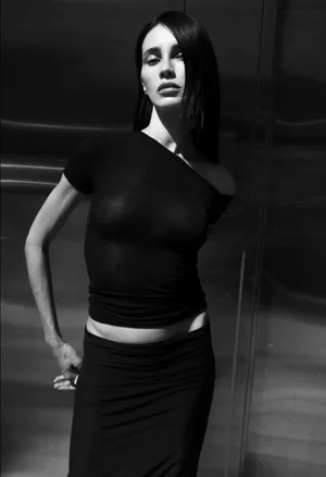 Black and white portrait of a woman wearing a dark fitted top and skirt against a dark textured background.