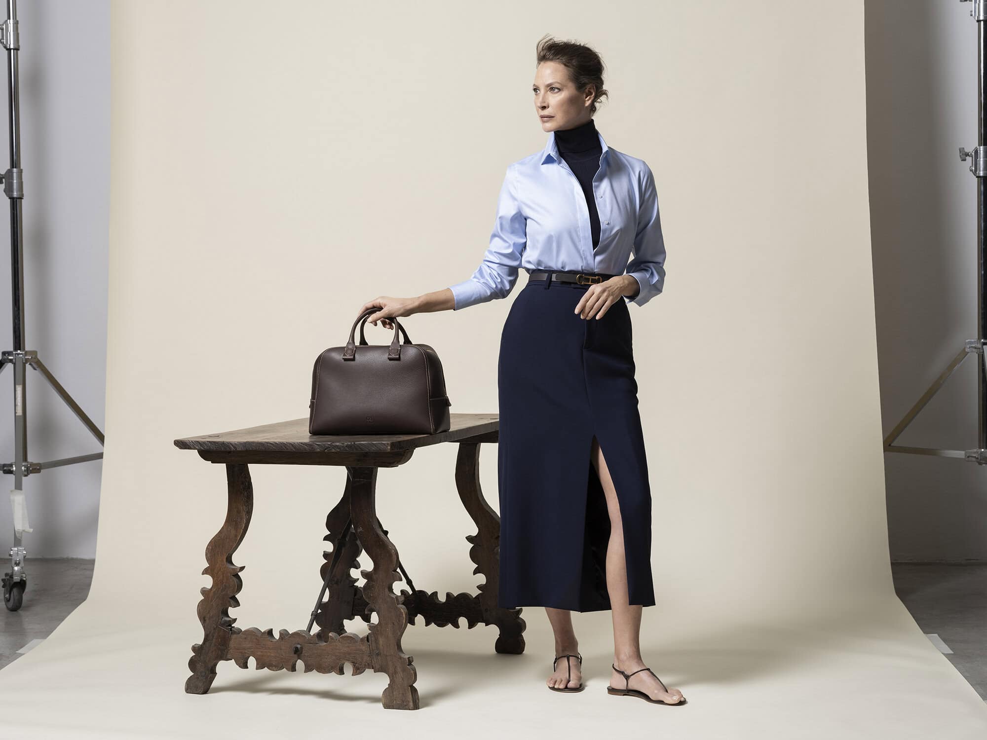 Elegant woman in a blue blouse and skirt poses in a studio, placing her hand on a brown leather handbag displayed on a wooden table.