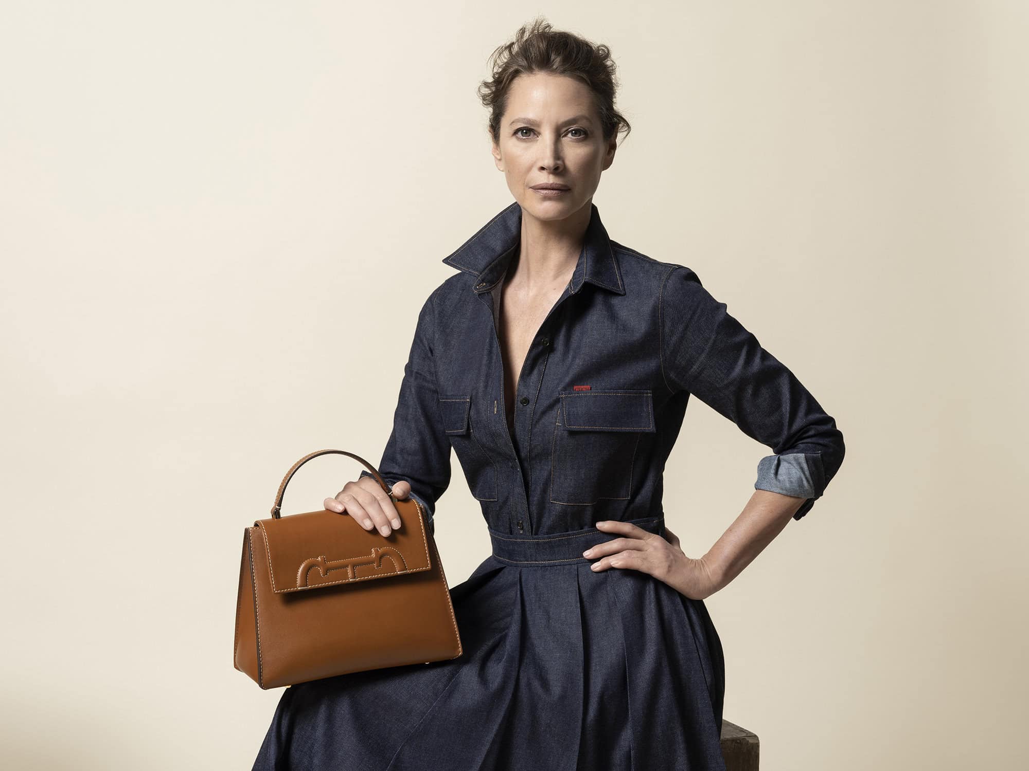 Adult female fashion model poses in a denim dress while holding a tan-colored handbag in a studio with a neutral-colored backdrop.