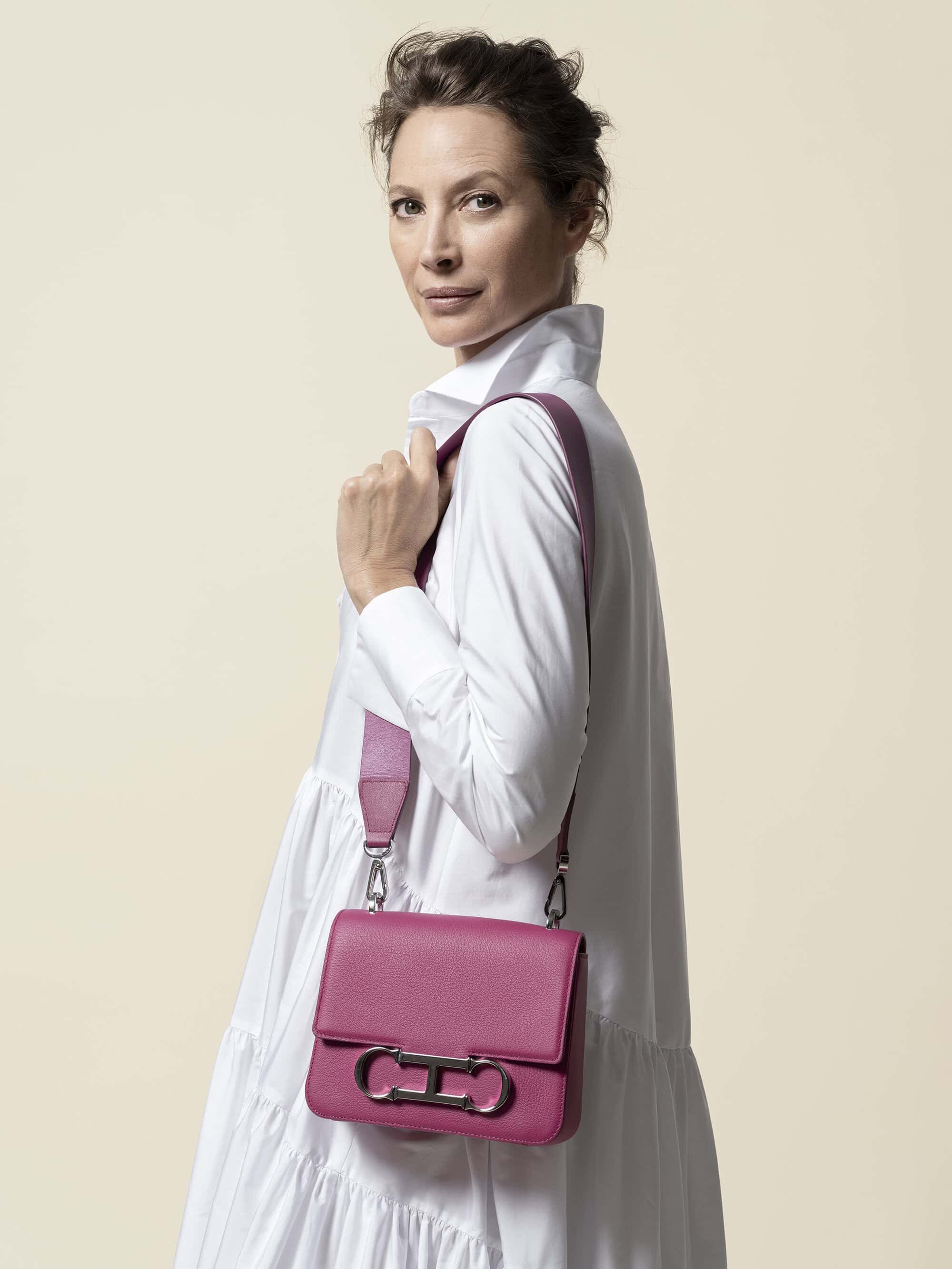 Brunette woman with a neutral expression models a pink leather crossbody bag with a silver clasp and a matching pink strap in a studio.