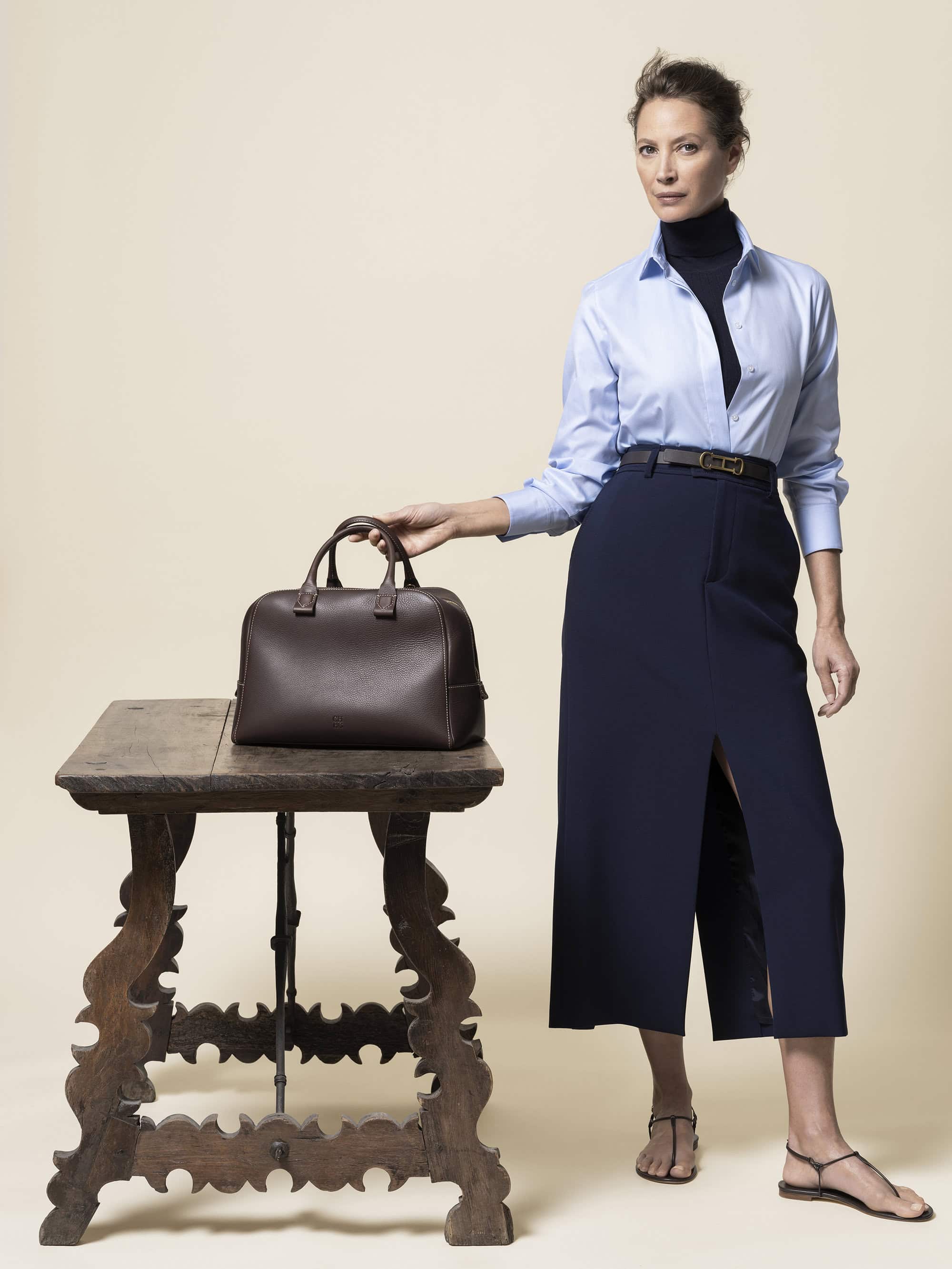 Woman posing with leather handbag on a wooden table. She is wearing a light blue shirt and navy skirt, set against a light beige background.