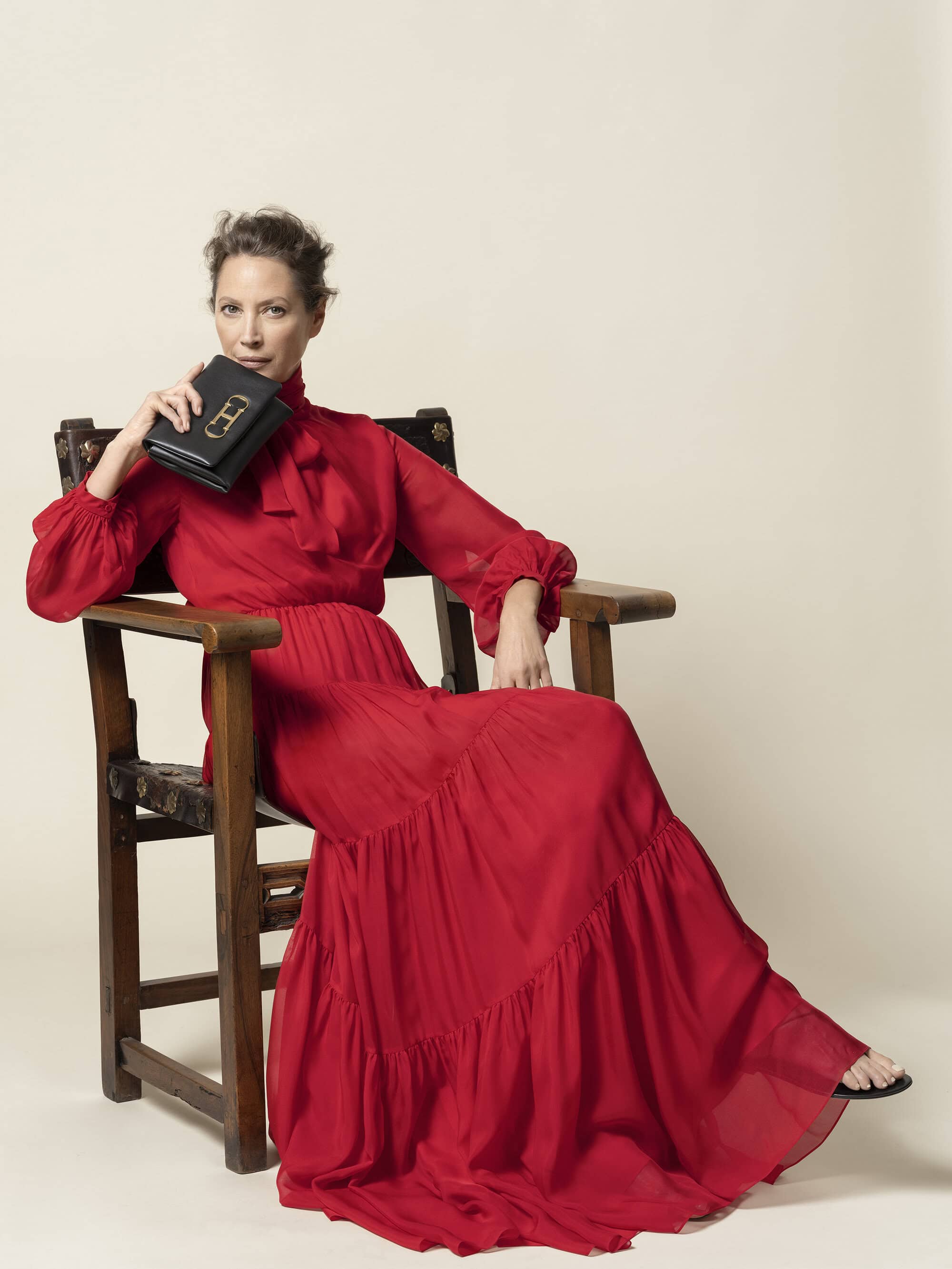 Elegant woman in a red chiffon gown sits in a wooden chair holding a black leather purse with a metal clasp in a photo studio.