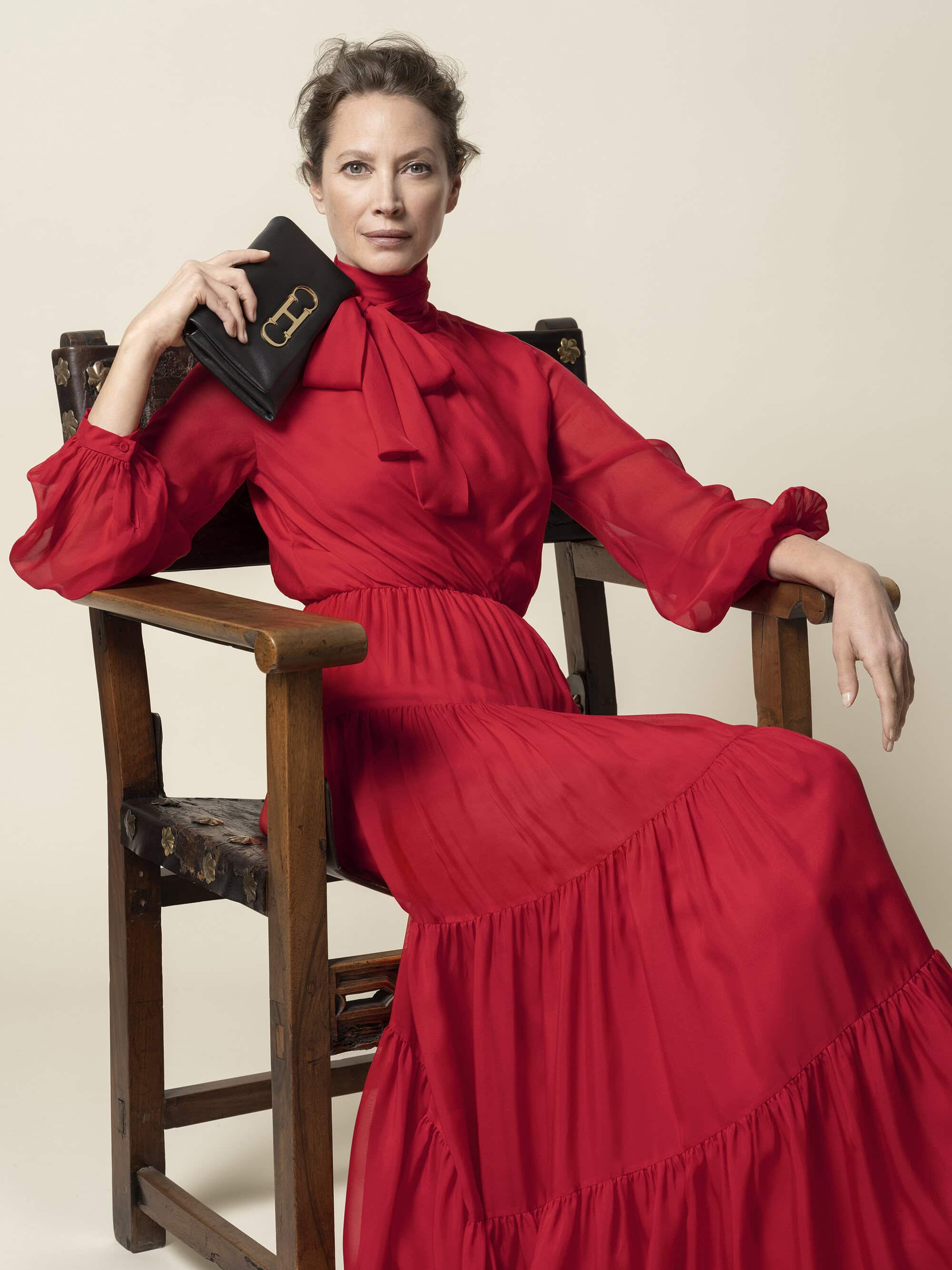 Adult female sitting in a chair wearing a red dress while holding a small black purse with a gold buckle, against a beige backdrop.