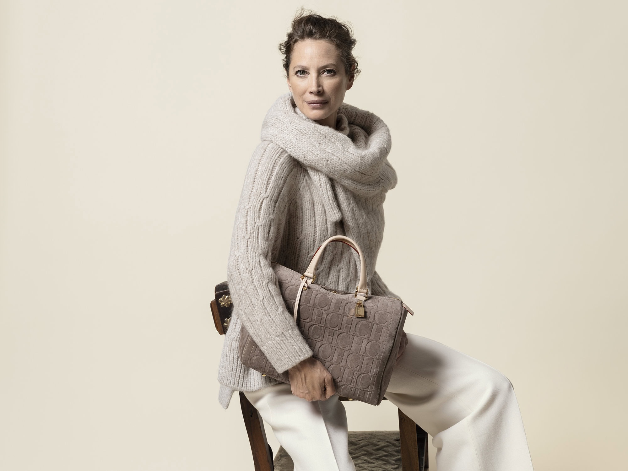 Adult woman in a cozy sweater sits and holds a beige handbag with brand logos; indoor neutral setting, soft lighting, fashion photography.