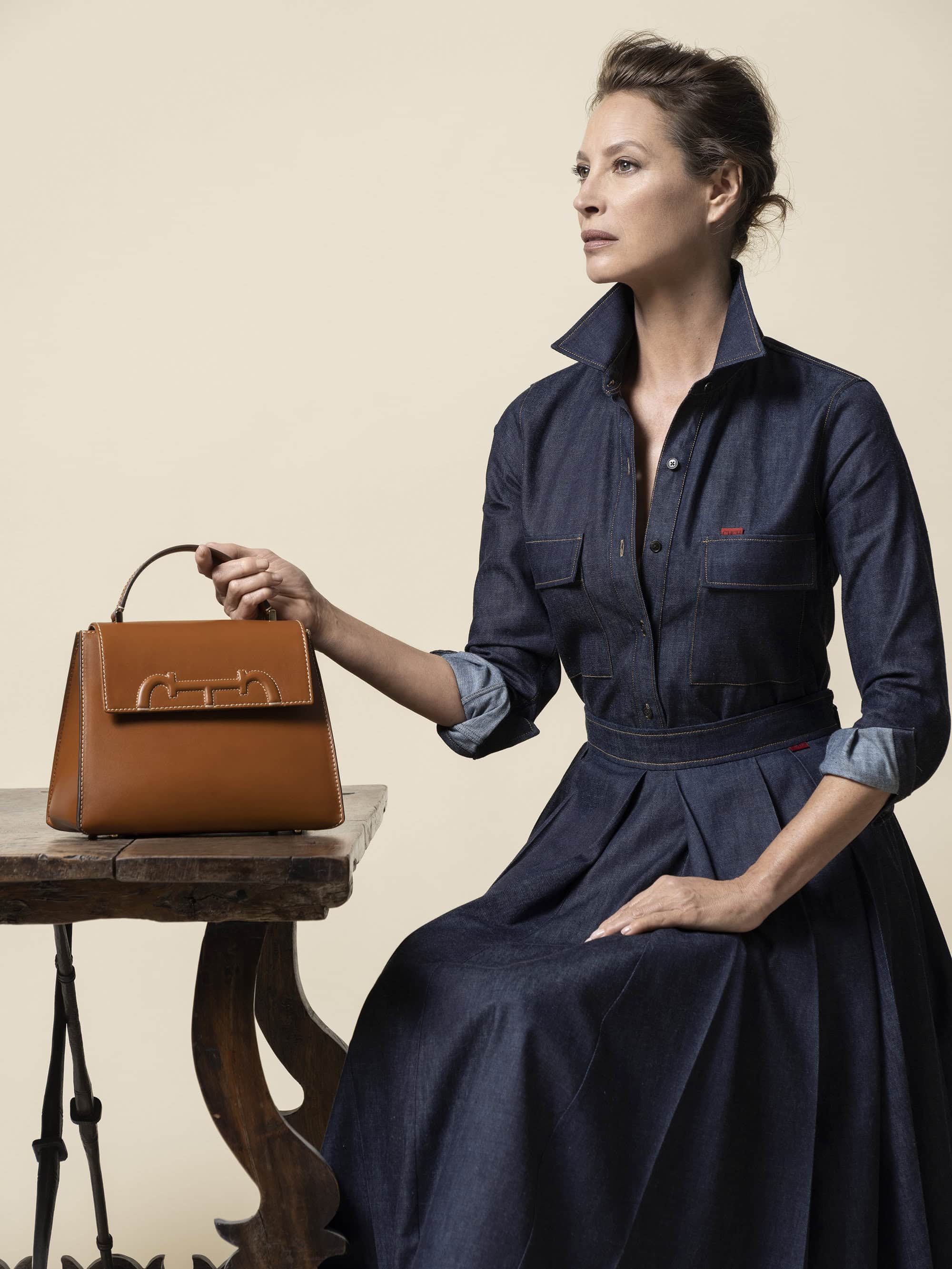 Woman in a denim dress sits next to a brown leather satchel bag on a wooden table against a beige background in a fashion setting.