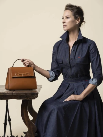 Woman in a denim dress sits next to a brown leather satchel bag on a wooden table against a beige background in a fashion setting.