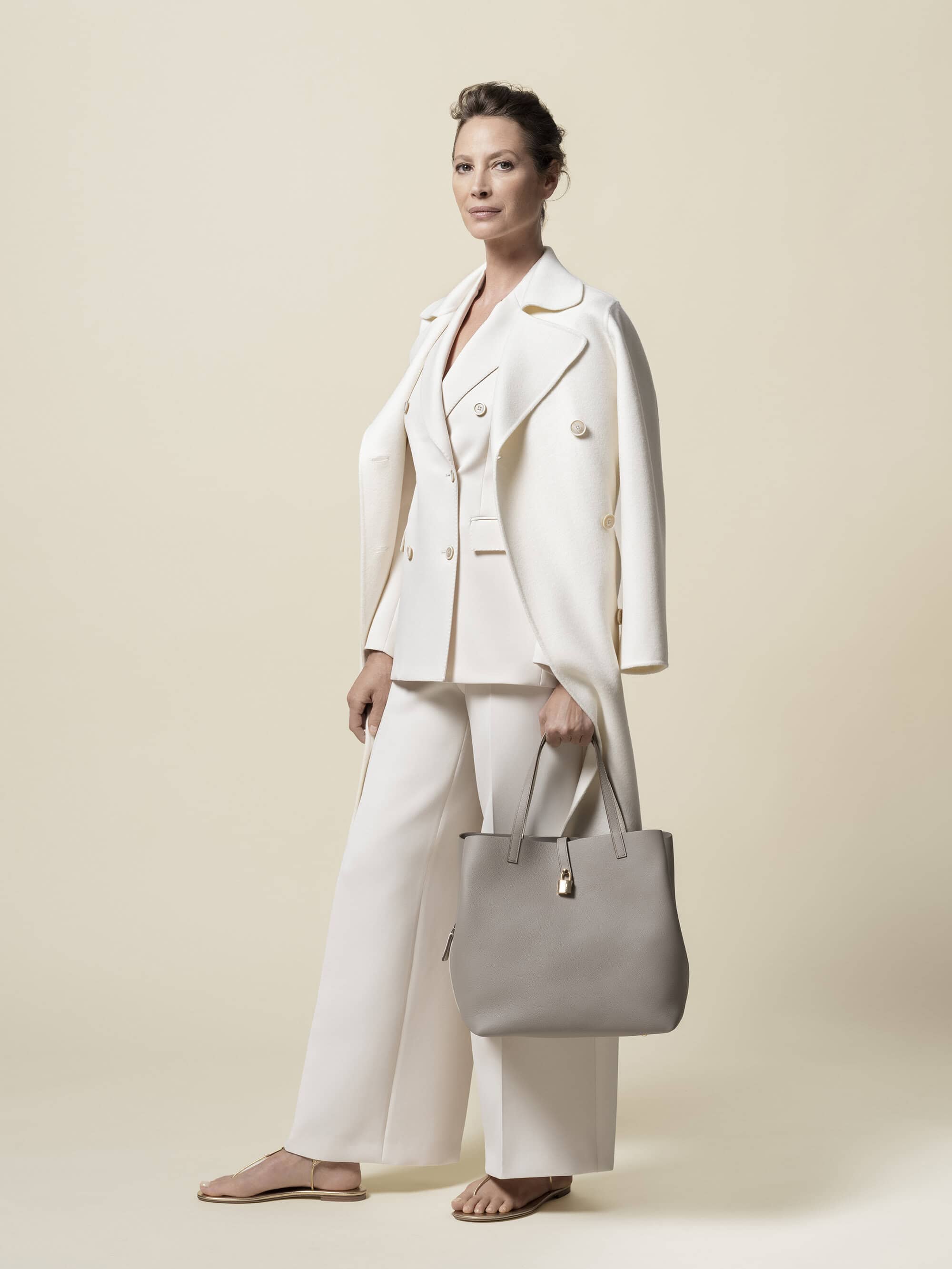 Woman poses in a studio wearing a white pantsuit and holding a handbag, standing against a neutral backdrop in a fashion editorial style.