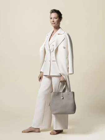Woman poses in a studio wearing a white pantsuit and holding a handbag, standing against a neutral backdrop in a fashion editorial style.