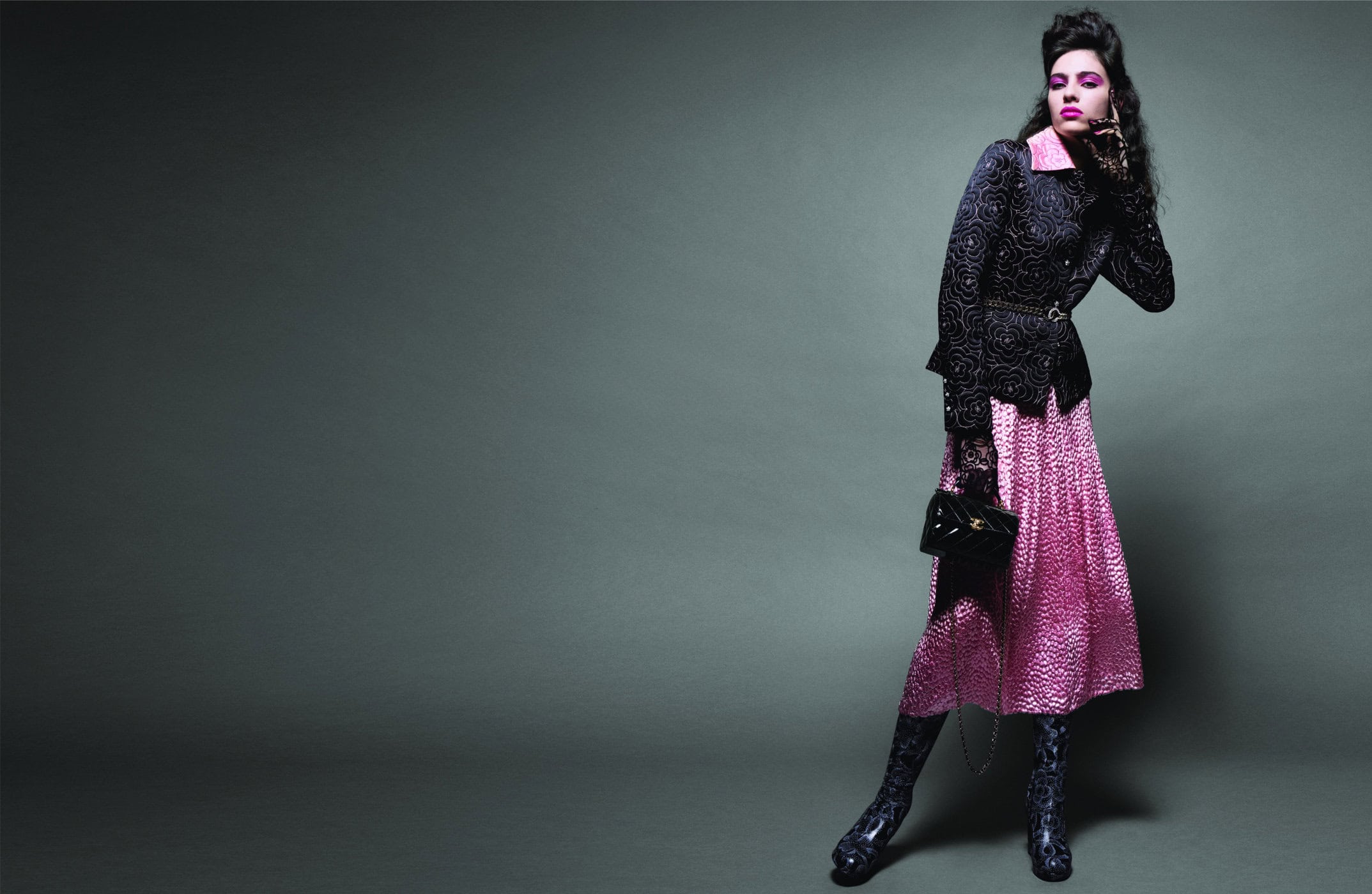 A fashion model poses in a black floral jacket and pink dress in a studio setting, complemented by black boots and a handbag.