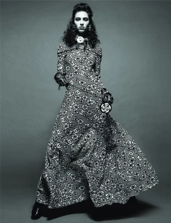 A model in a long, patterned Chanel dress poses in a studio with a neutral background, holding a circular floral-patterned purse.