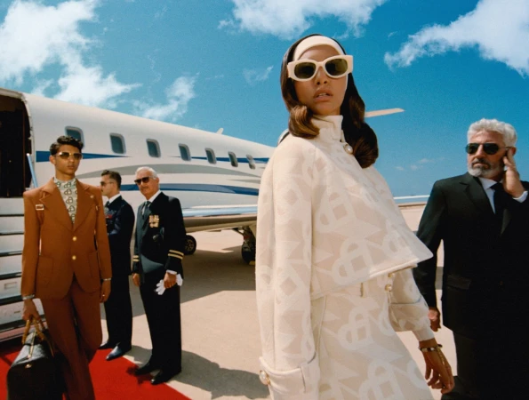 Fashionable woman in sunglasses and patterned outfit stands near a private jet with suited men, under a bright sky, runway setting.