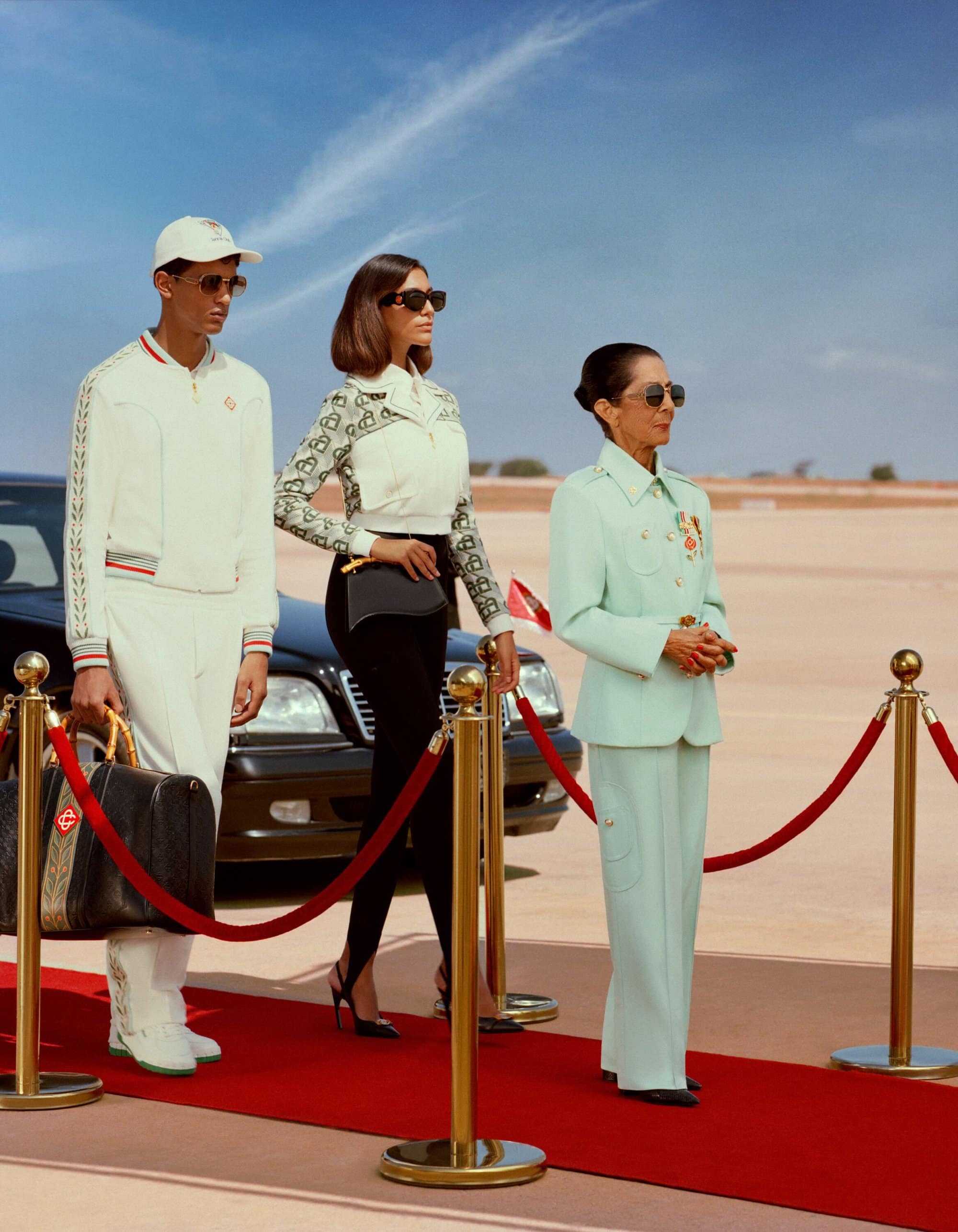 Three fashionably dressed people on a red carpet with a luxury car, showcasing an outdoor editorial fashion shoot under a bright blue sky.