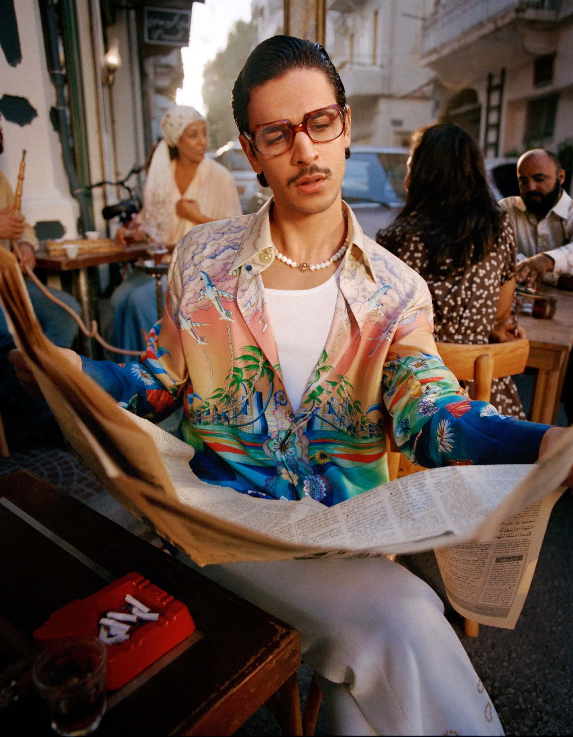 Man in patterned shirt reads newspaper at outdoor cafe. Several people sit at tables in the background on a sunny day.