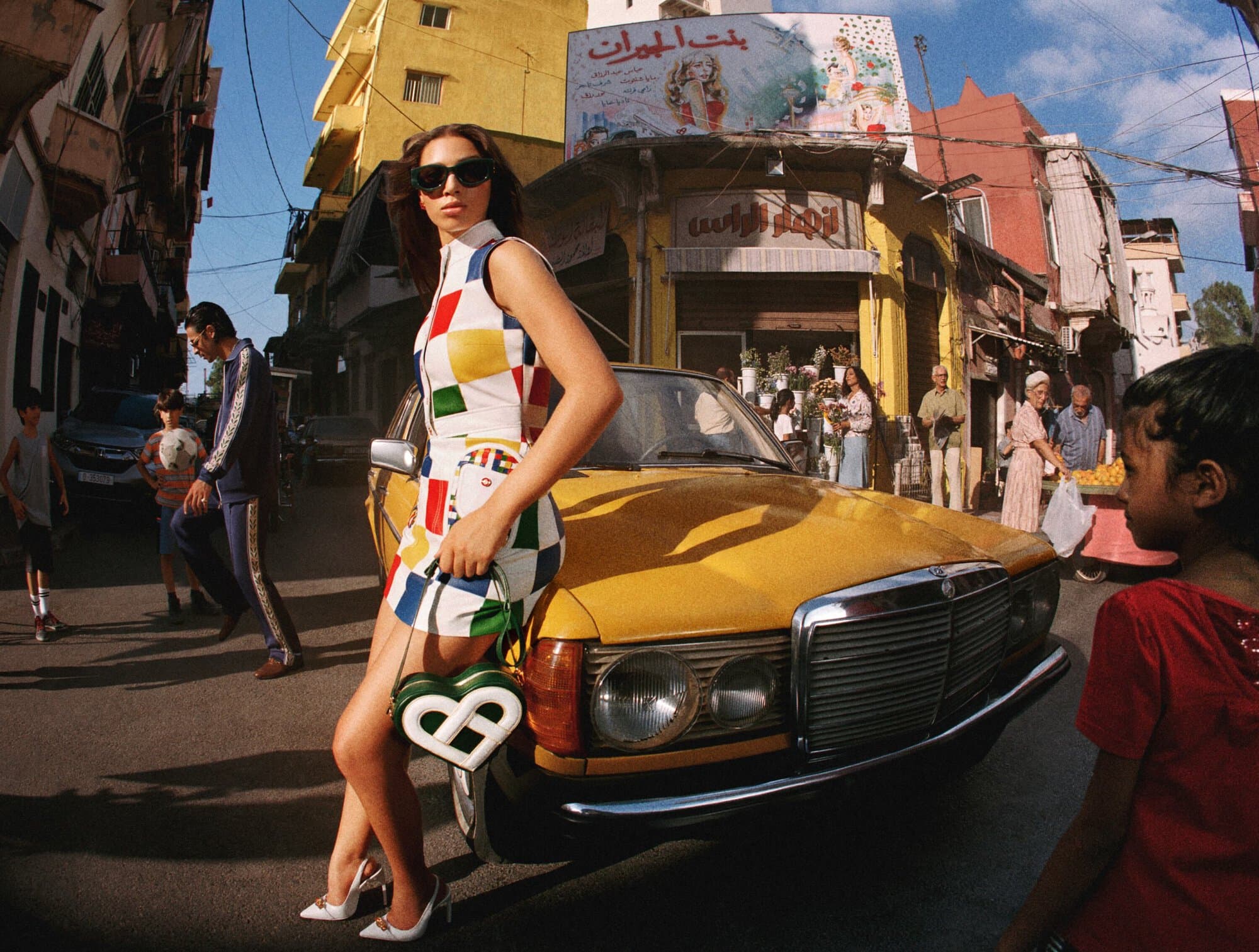 Fashionable woman in checkered mini dress poses next to a yellow vintage car on a city street with pedestrians and buildings.