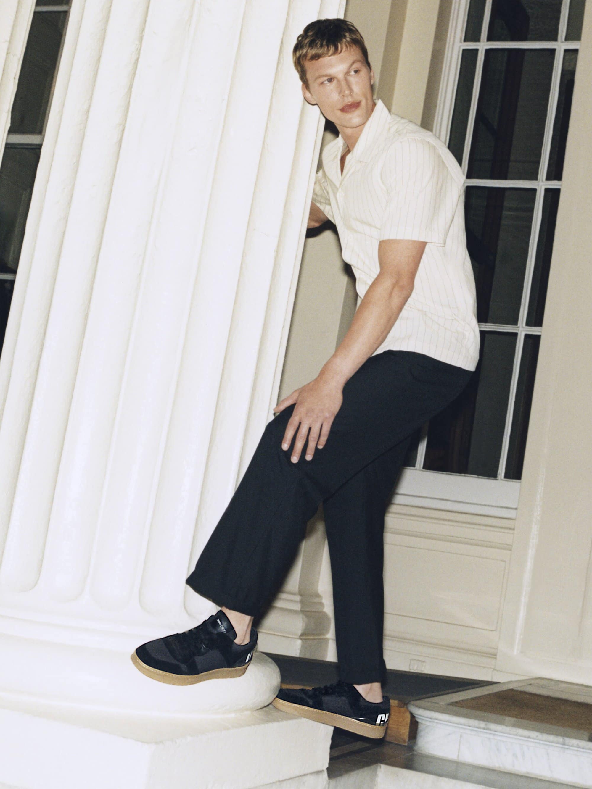 Blonde man in a cream-colored shirt and black pants poses with black sneakers next to a white pillar indoors.
