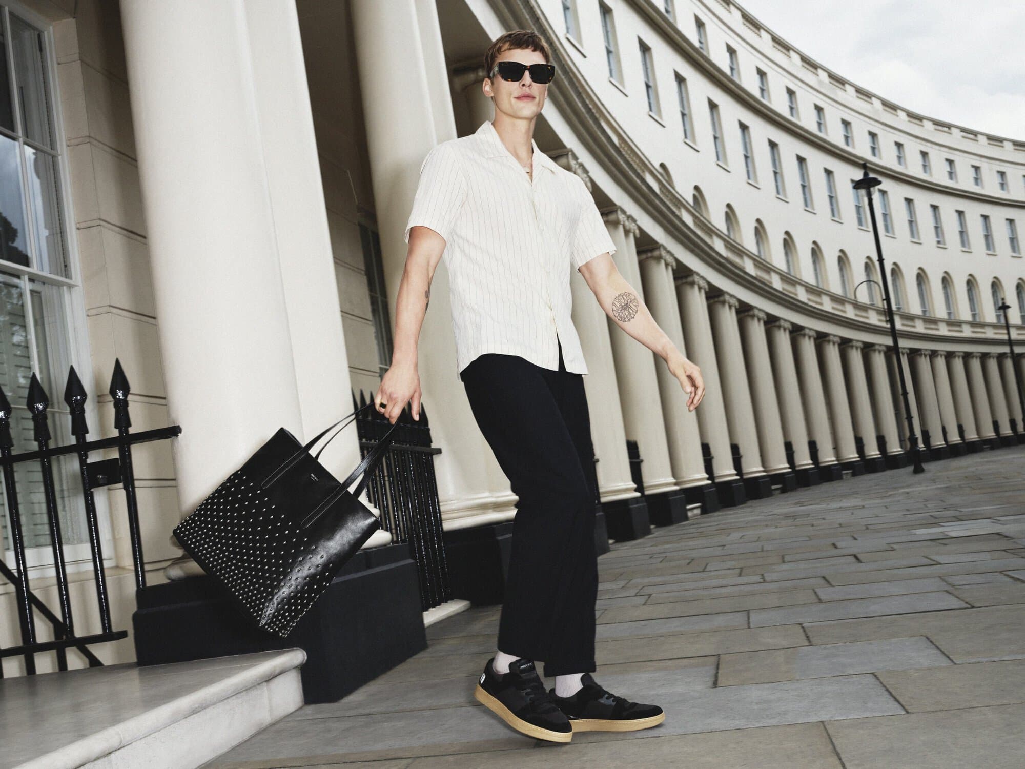 A young adult man walks down a city street in sunglasses, carrying a studded black tote bag, with classic buildings in the background.