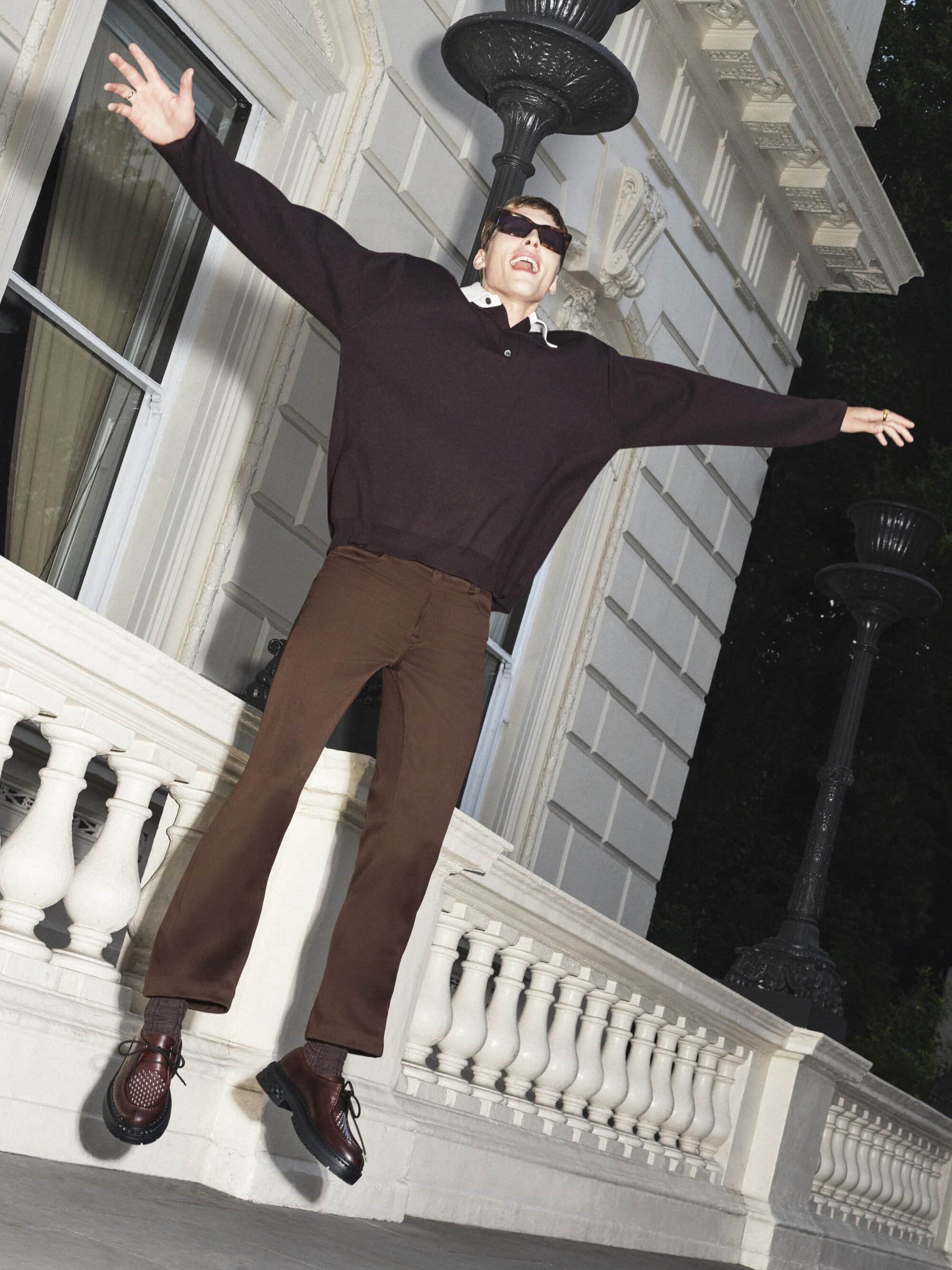Man in brown sweater and pants leaps outdoors on a city street against a light-colored building, smiling with arms outstretched.
