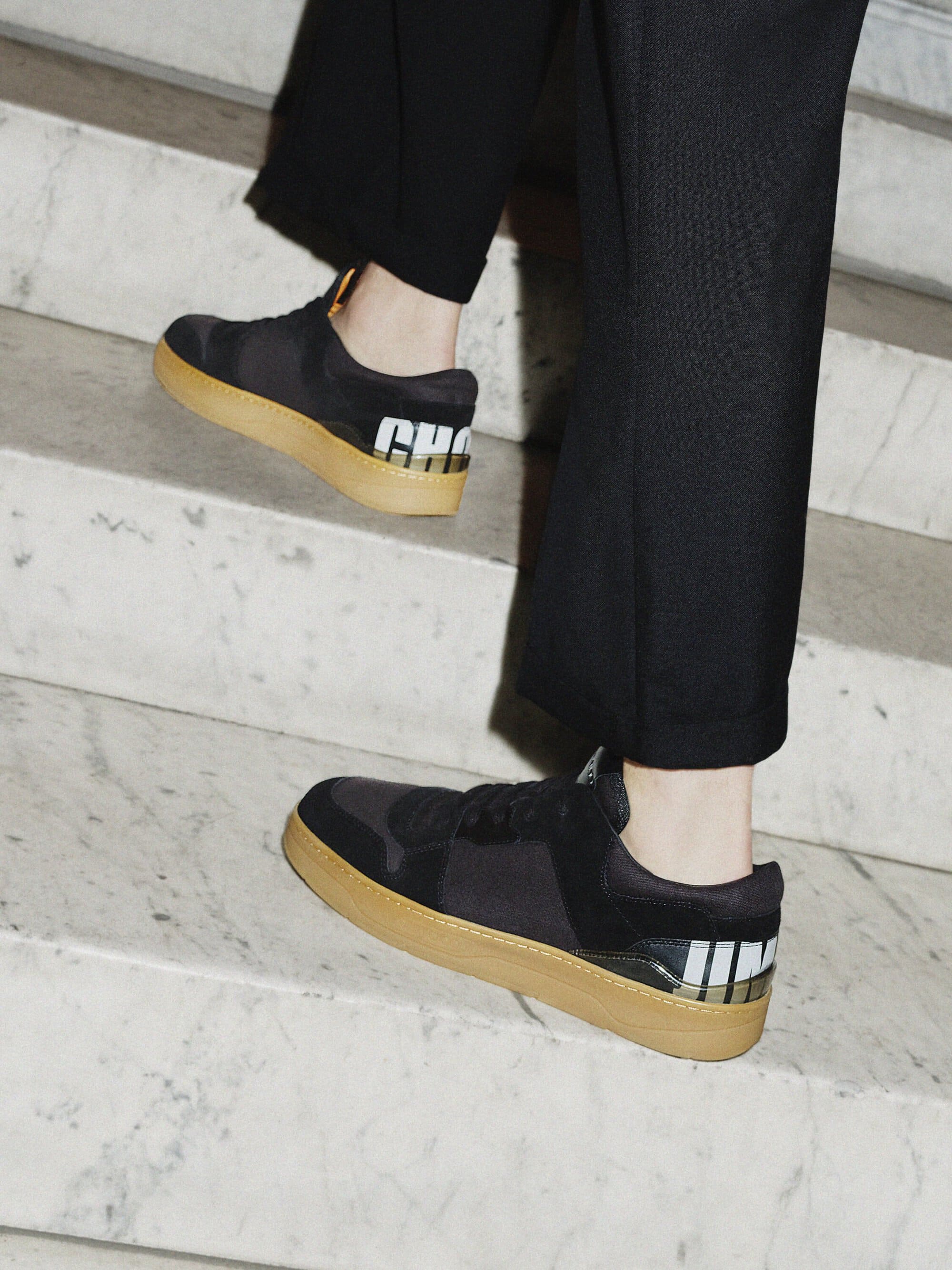 A pair of black sneakers with tan soles is shown on marble steps, along with dark-colored pants and a partial view of a person's legs.