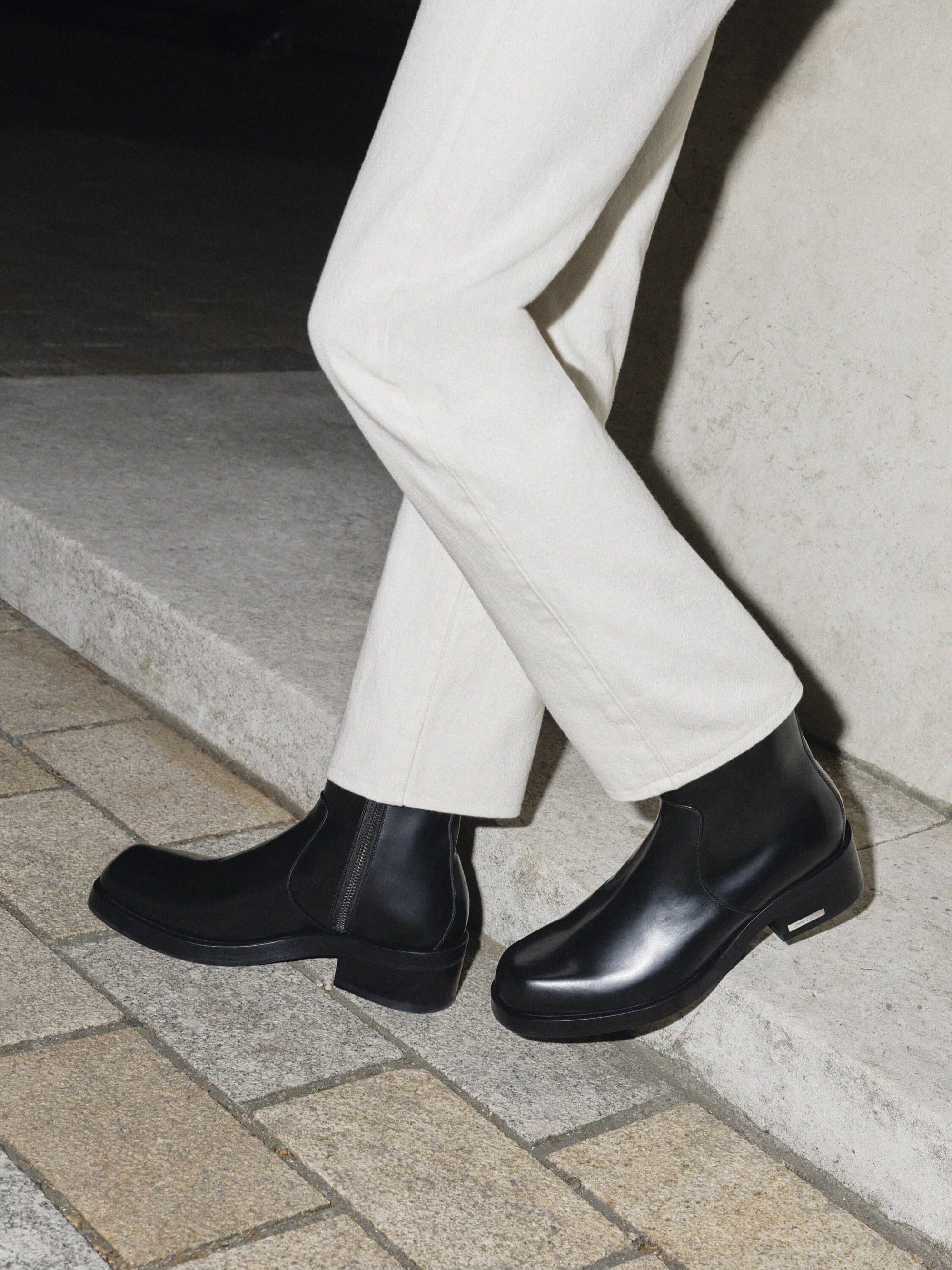 Close-up of black leather Chelsea boots with elastic side panels and white denim jeans against a neutral-toned architectural backdrop.