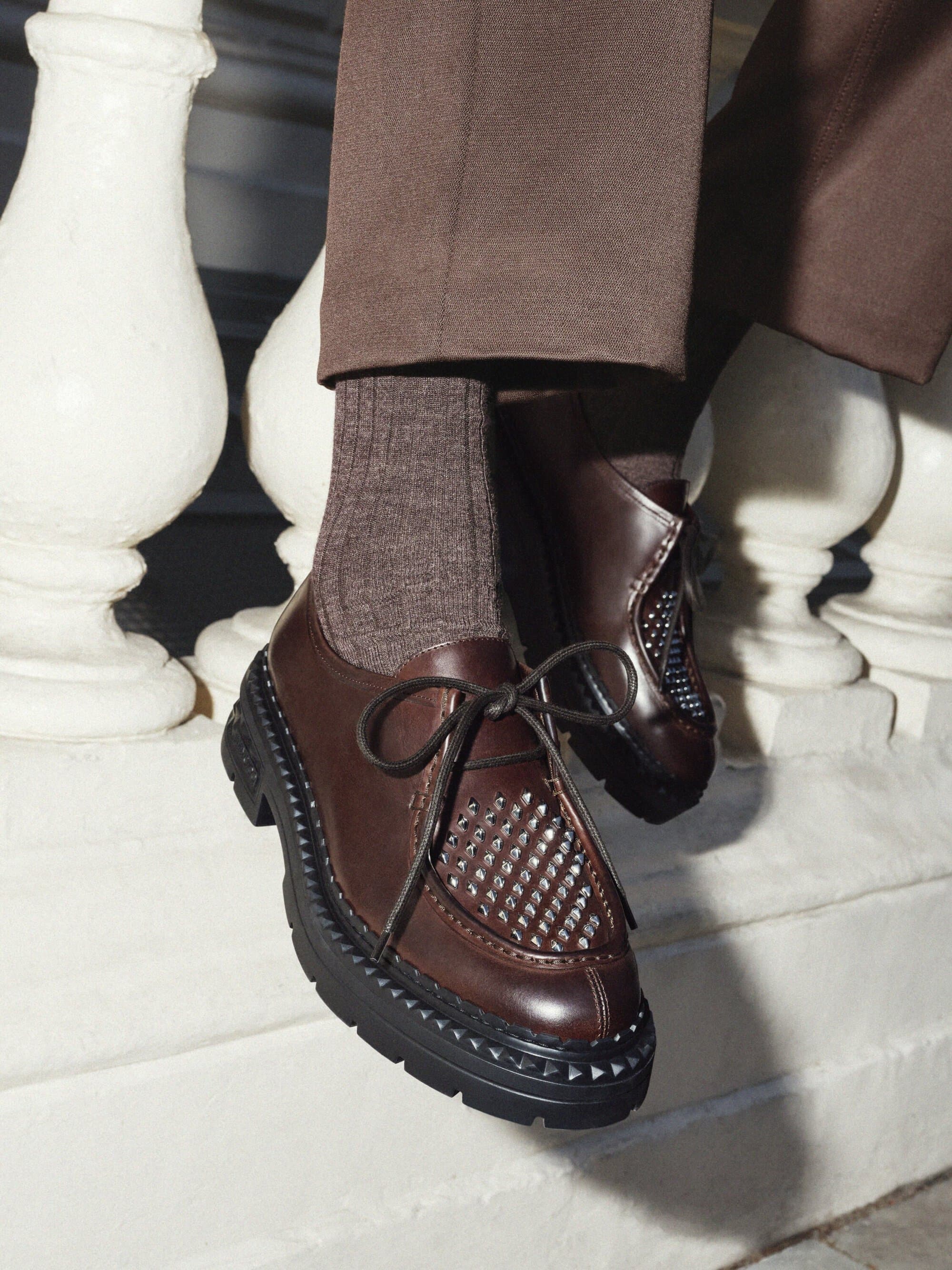 Close-up of dark brown leather dress shoes with a thick black lug sole, brown socks, and brown trousers on a white ledge.