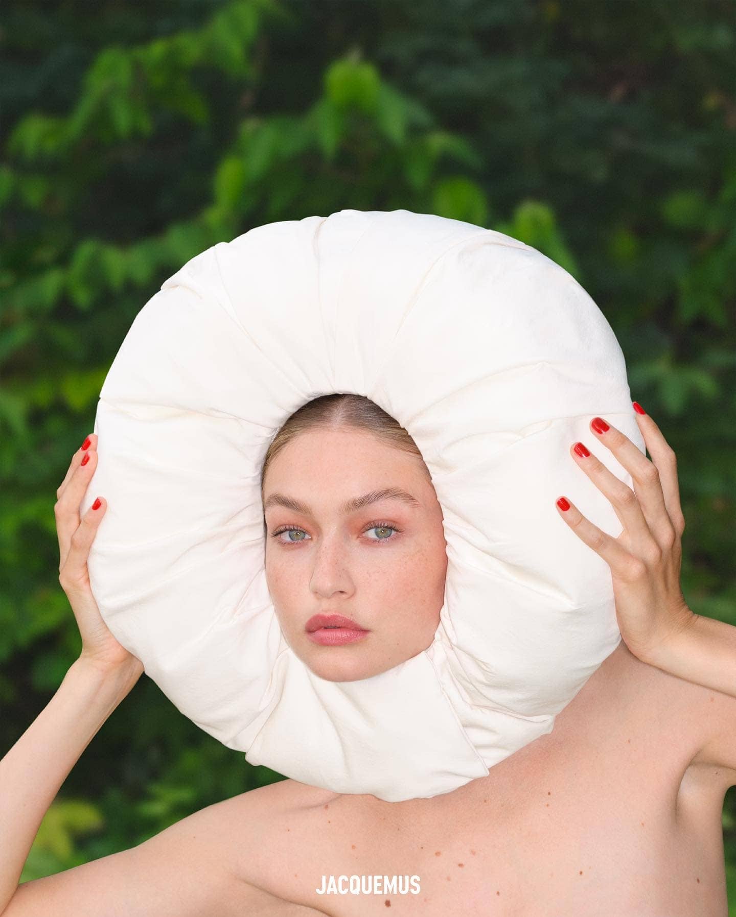 Fashion portrait of a woman with red nails, wearing a large white round headpiece against a leafy green background with the Jacquemus logo.