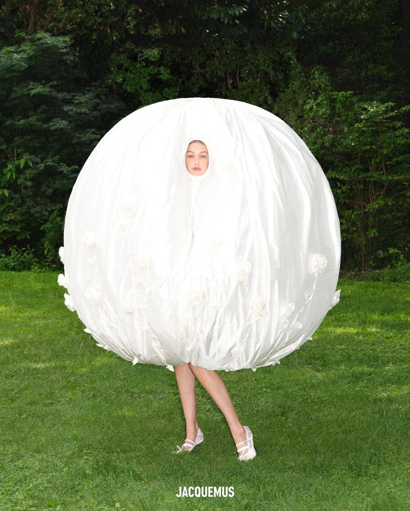 A fair-skinned adult female poses in a large round white garment in an outdoor setting with a green grassy lawn and trees in the background.
