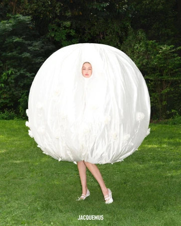 A fair-skinned adult female poses in a large round white garment in an outdoor setting with a green grassy lawn and trees in the background.