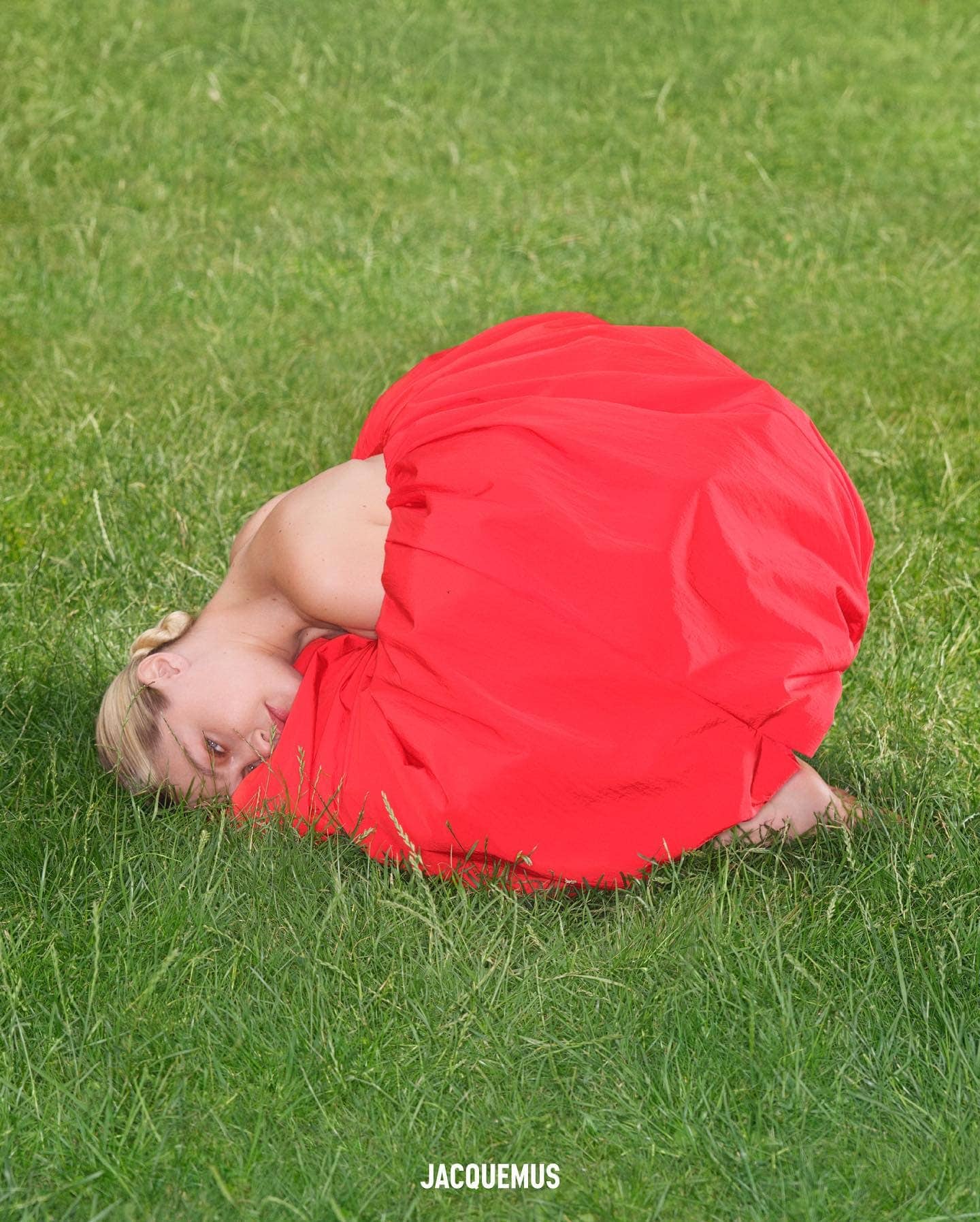 Woman in a red dress lying in green grass in a fashion editorial shot. The model poses outside in bright sunlight with a blurred background.