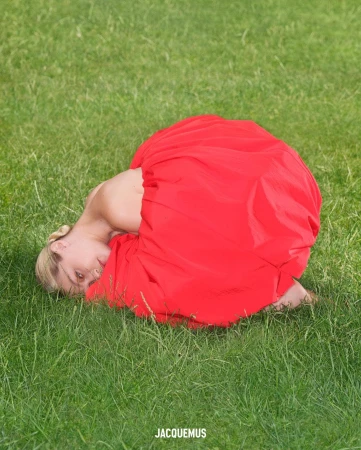 Woman in a red dress lying in green grass in a fashion editorial shot. The model poses outside in bright sunlight with a blurred background.