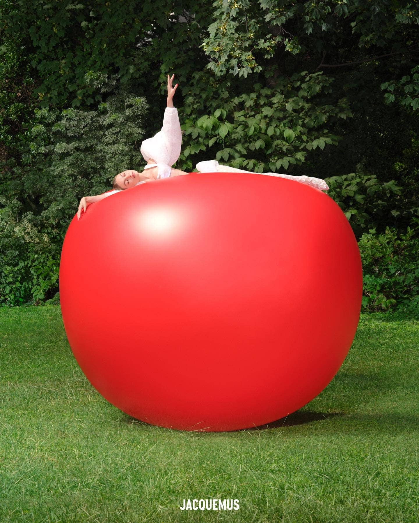 Adult woman in white apparel posing atop a giant red sphere on grass, with a lush green forest as backdrop and brand logo 'Jacquemus' visible.