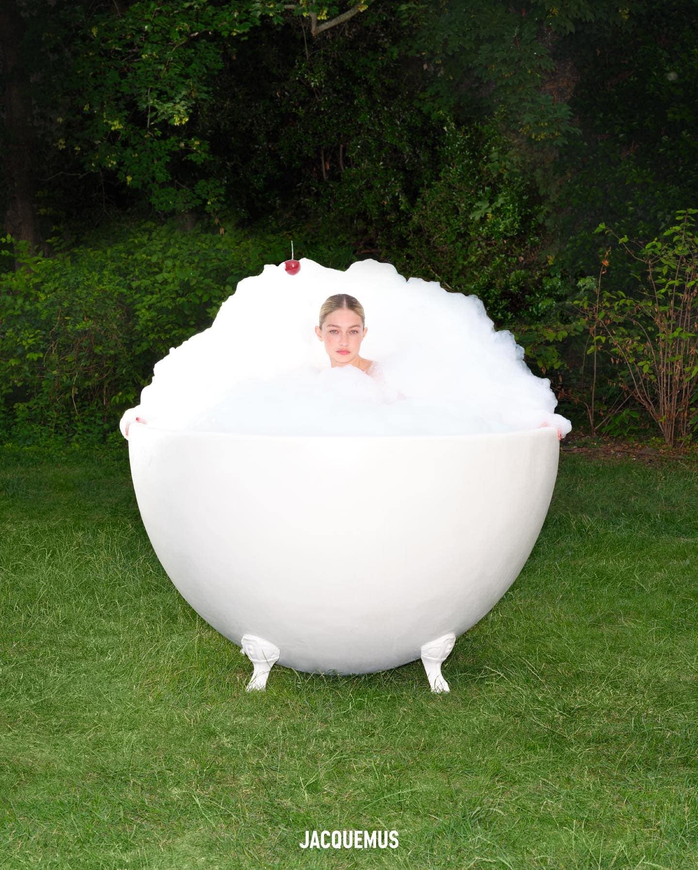 An adult woman sits in a white bathtub filled with bubbles outdoors surrounded by green trees and grass in a beauty or fashion shot.