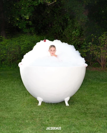 An adult woman sits in a white bathtub filled with bubbles outdoors surrounded by green trees and grass in a beauty or fashion shot.
