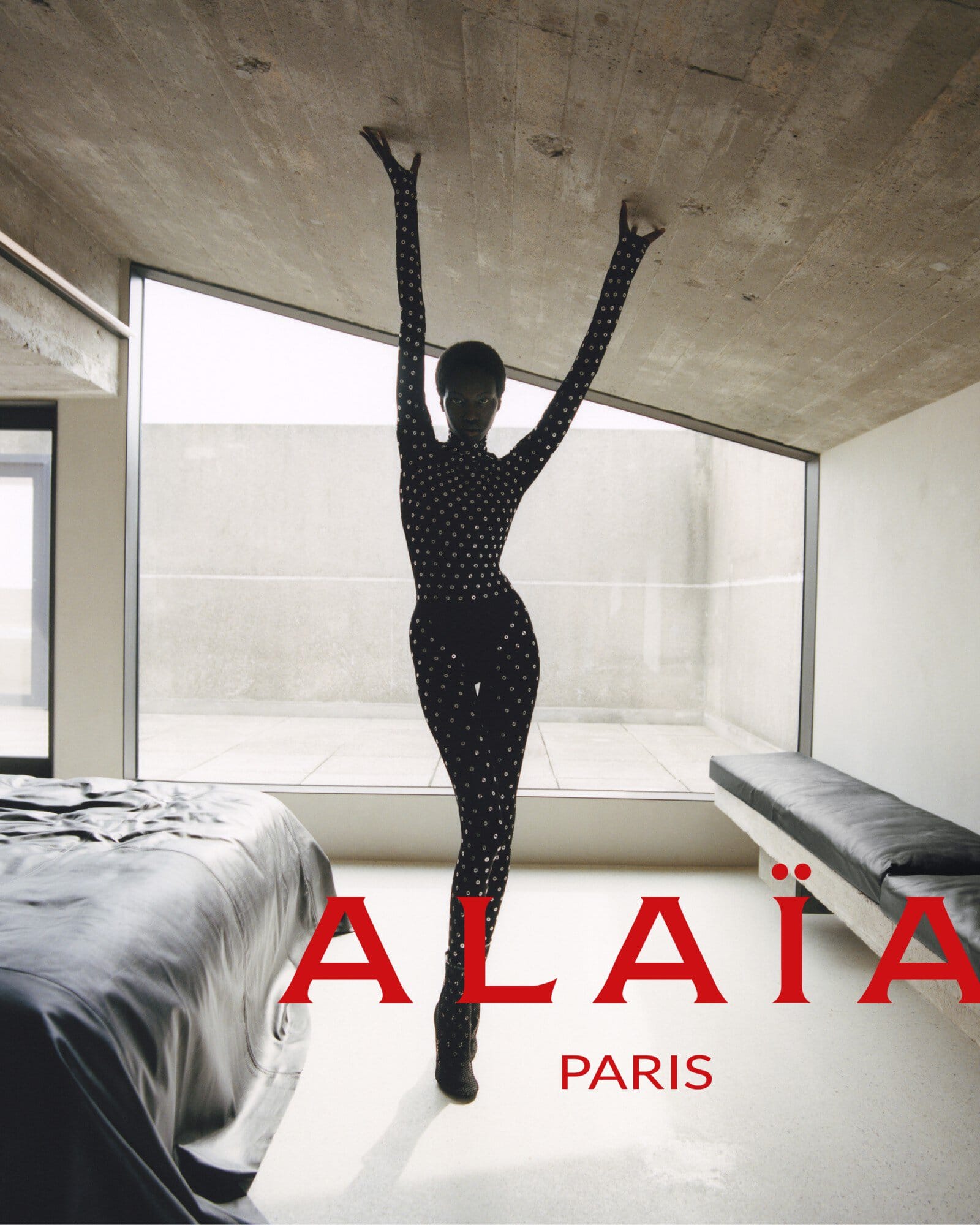 Fashion editorial shot of a Black female model in a black studded bodysuit, striking a pose indoors against a modern architectural background.