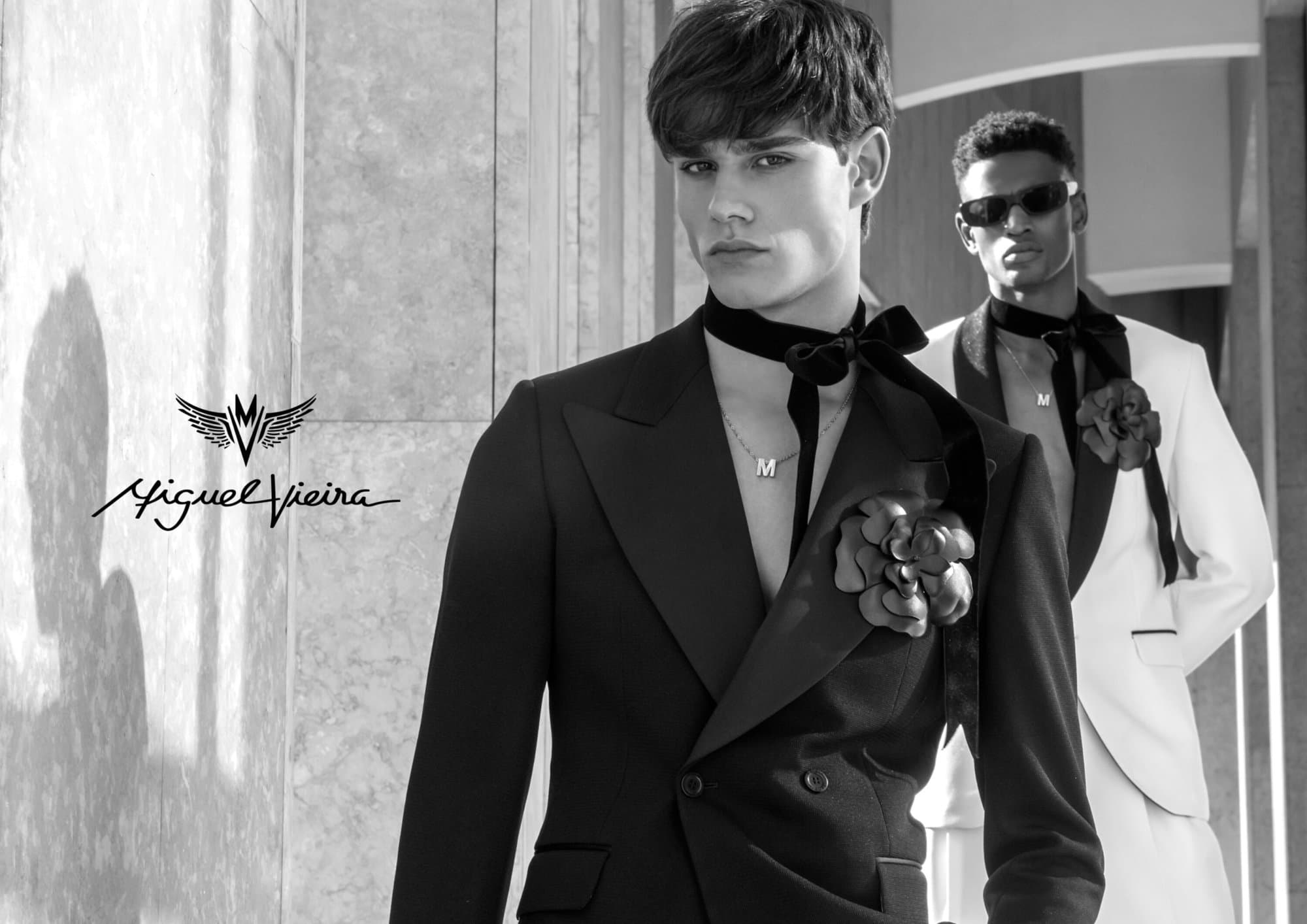 Two men in formal black and white suits with velvet scarves and floral brooches, posing indoors with soft window lighting. Fashion.