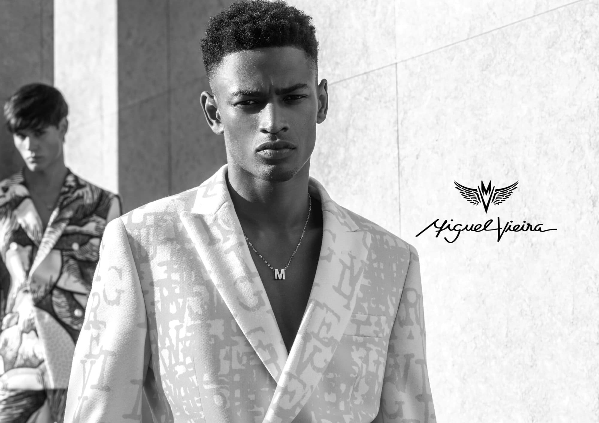 Monochrome shot of two adult male models wearing patterned blazers in front of a light grey wall with visible logo of Miguel Vieira.