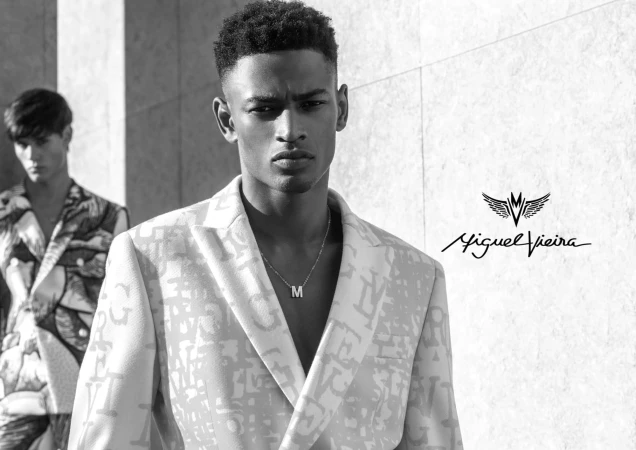 Monochrome shot of two adult male models wearing patterned blazers in front of a light grey wall with visible logo of Miguel Vieira.