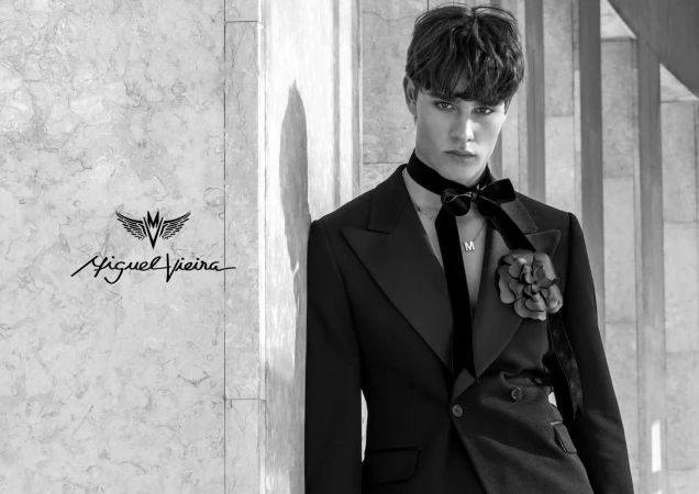 Monochrome portrait of a young adult male model in a dark suit, posing against a textured wall with a fashion logo, in a studio setting.