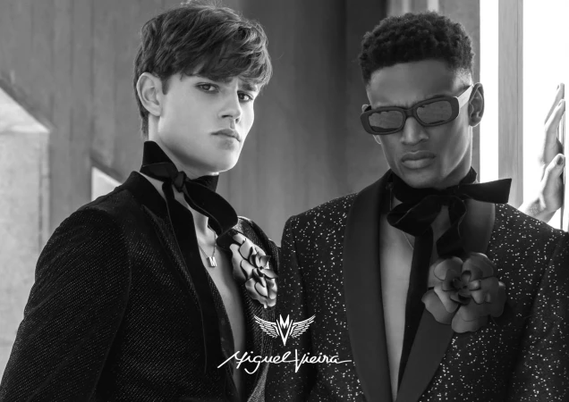 Two stylish adult male models pose in black and white, wearing tuxedo jackets, black ascots, and decorative flower brooches indoors.