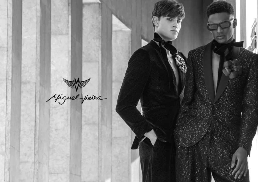 Two men in fashionable black suits stand side-by-side against a modern architectural backdrop, showcasing sophisticated formal wear.