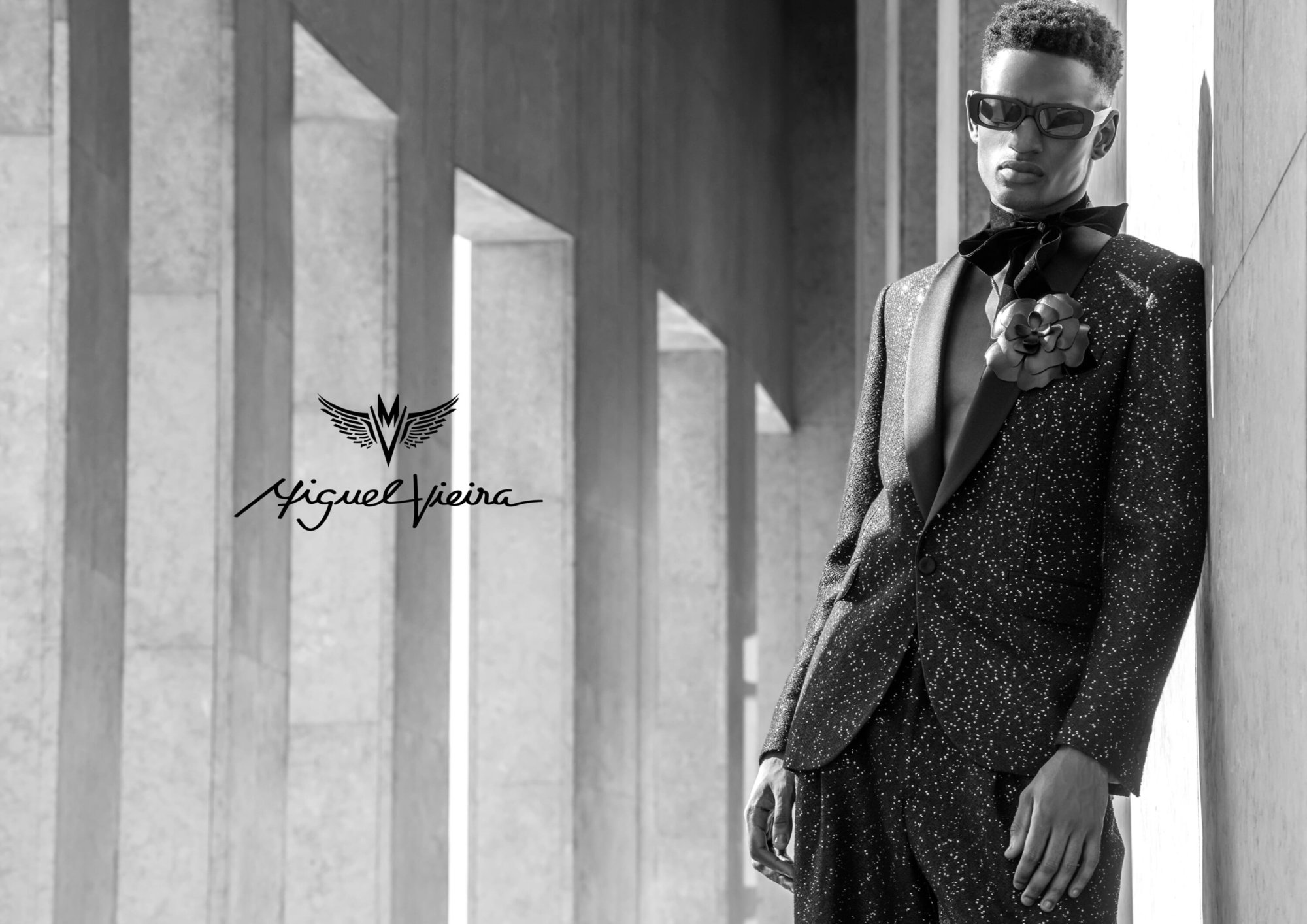 Black and white fashion portrait of a stylish Black male model in a tuxedo with a floral accessory, posing against a concrete wall.