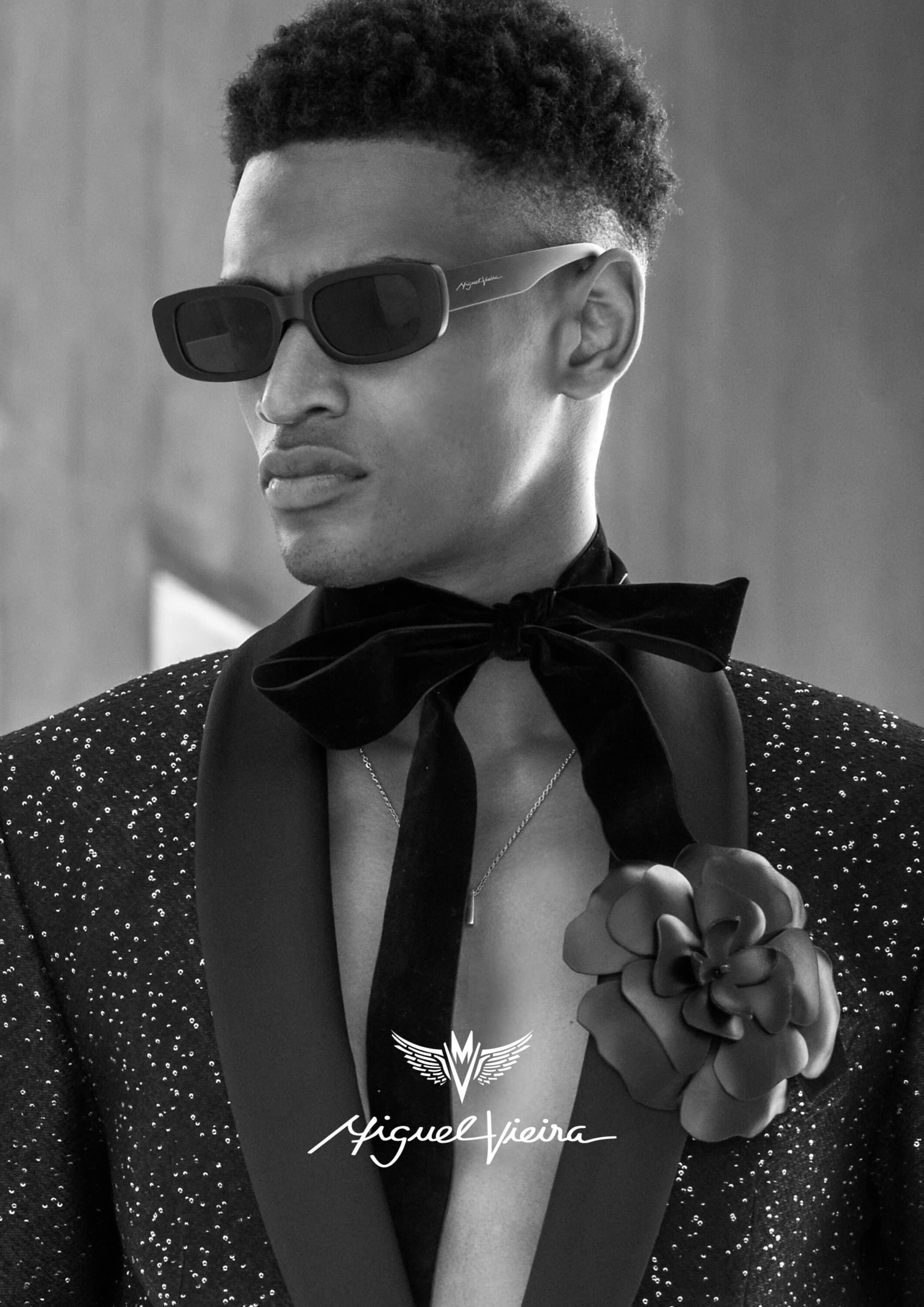 Close-up black and white portrait of a stylish man wearing sunglasses, a flower-pinned suit, and a velvet bowtie in a studio setting.