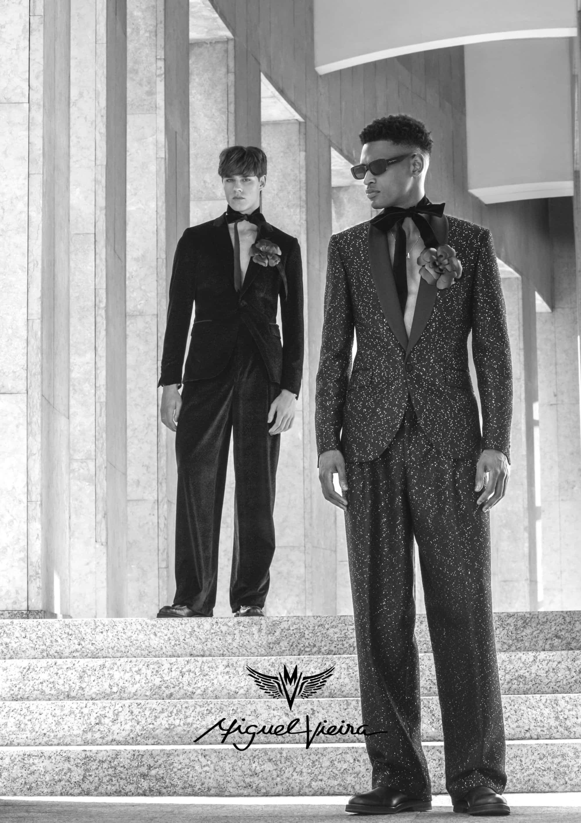 Two elegant male models showcase formal wear on a stone staircase, featuring suits with floral details in a monochromatic setting.